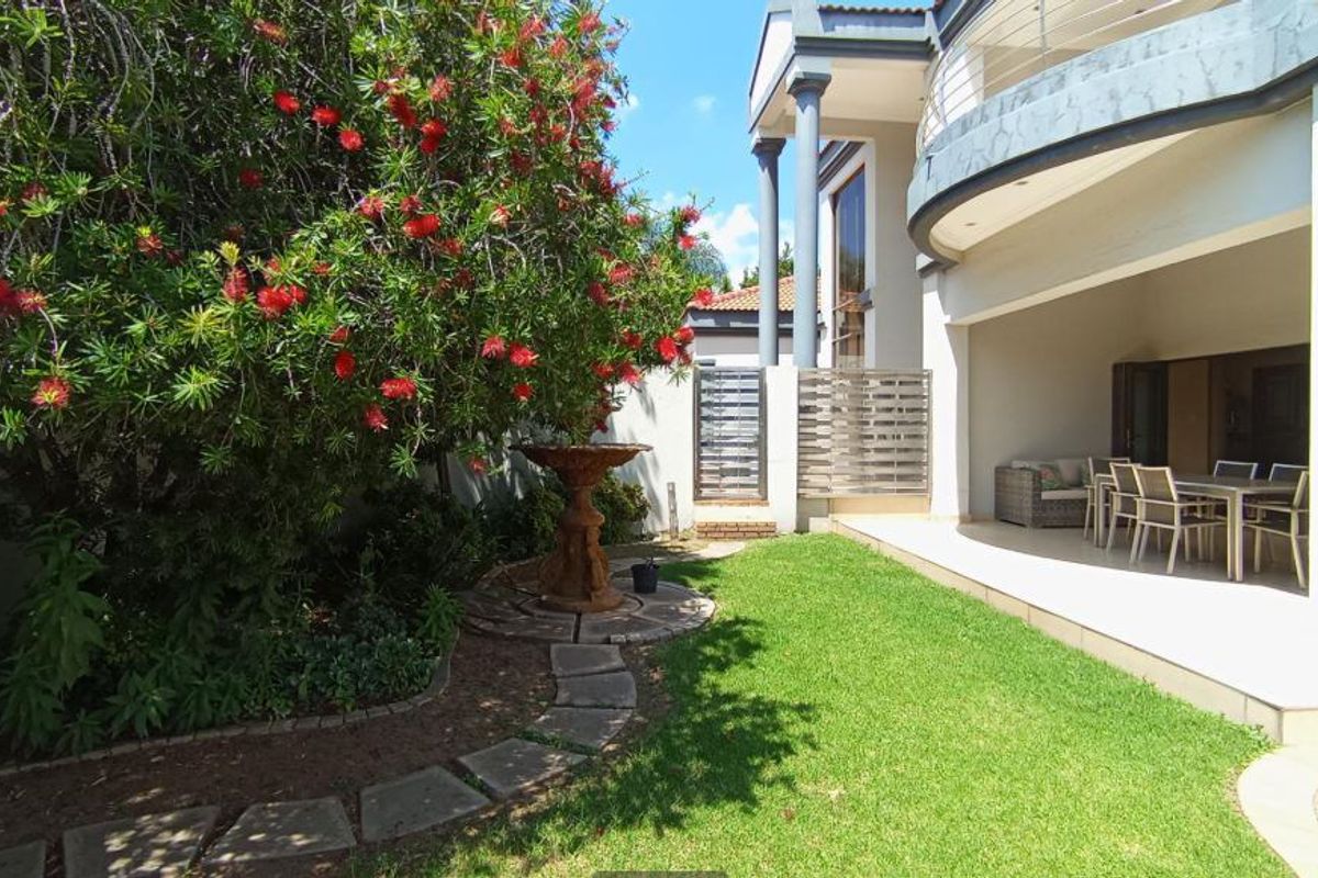 A well-maintained rear garden featuring a stone fountain, paved pathway, and a spacious covered patio area with access to the upper balcony.