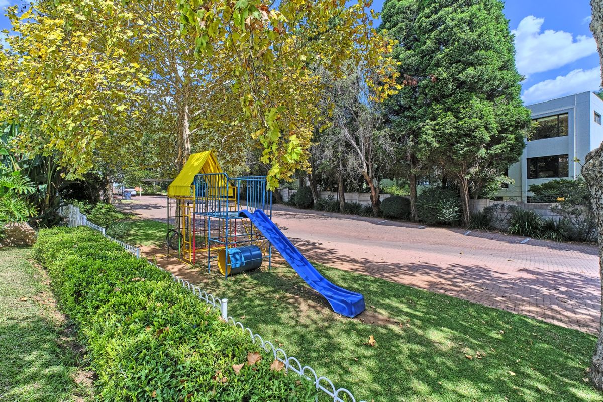 A well-maintained communal garden area featuring a paved driveway, mature trees, and a children's play structure within an apartment complex setting.