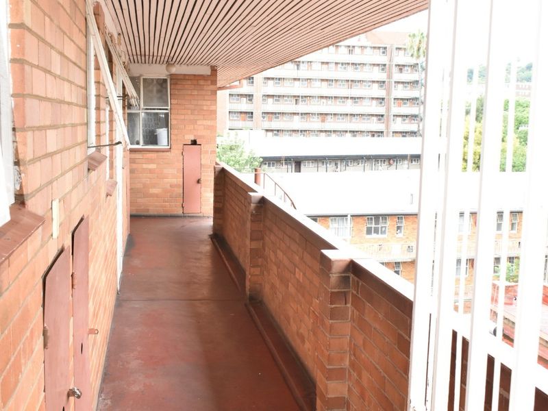 An exterior balcony featuring brick walls, a slatted ceiling, and a security gate, providing an outdoor walkway area for the apartment.