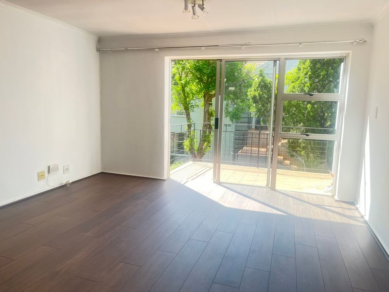 An empty living room featuring dark laminate flooring and a large sliding glass door that opens onto a private balcony with leafy views.
