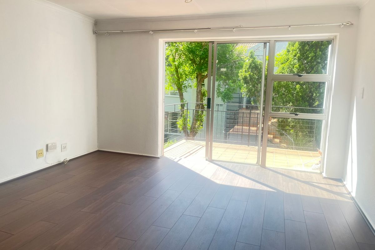 An empty living room featuring dark laminate flooring and a large sliding glass door that opens onto a private balcony with leafy views.