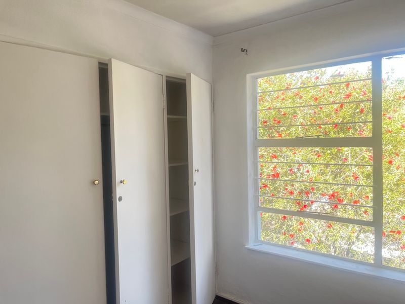 A bright, empty bedroom featuring built-in cupboards and a window with security bars overlooking a flowering tree.