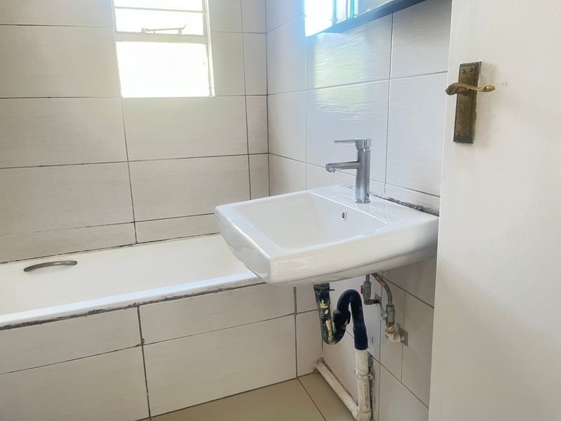 A functional bathroom featuring a wall-mounted basin, bathtub, and tiled finishes, currently requiring maintenance on plumbing and sealant.