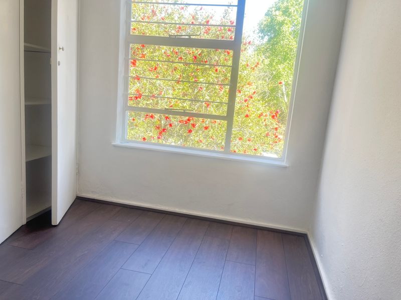 A bright, empty bedroom featuring dark laminate flooring, a built-in cupboard, and a large window with security bars overlooking greenery.