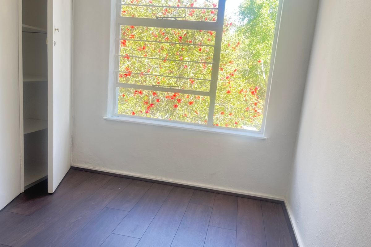A bright, empty bedroom featuring dark laminate flooring, a built-in cupboard, and a large window with security bars overlooking greenery.