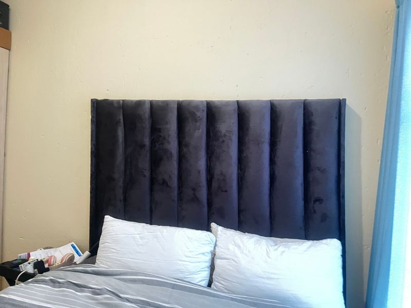 A bedroom interior featuring a dark upholstered headboard against a plain wall with visible plaster imperfections.