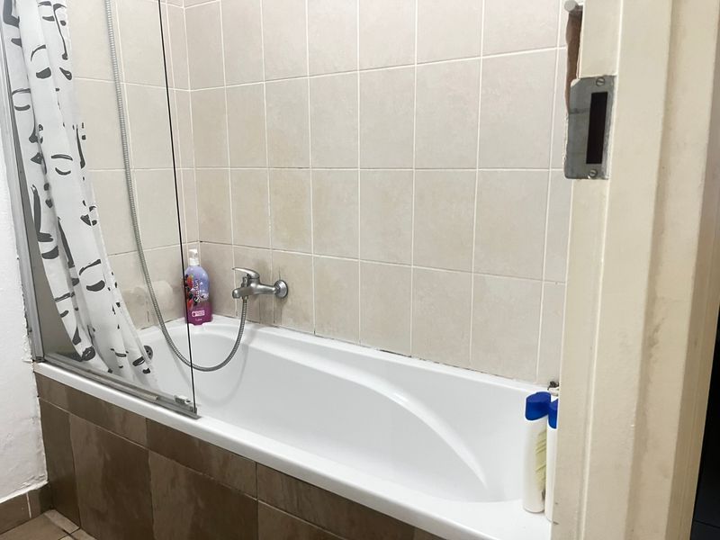 A bathroom featuring a bathtub with a wall-mounted shower mixer, glass screen, and tiled surround, showing signs of wear and dated finishes.