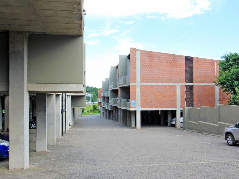 Exterior view of an apartment complex featuring exposed brick architecture, concrete structural pillars, and a paved driveway with covered parking spaces.