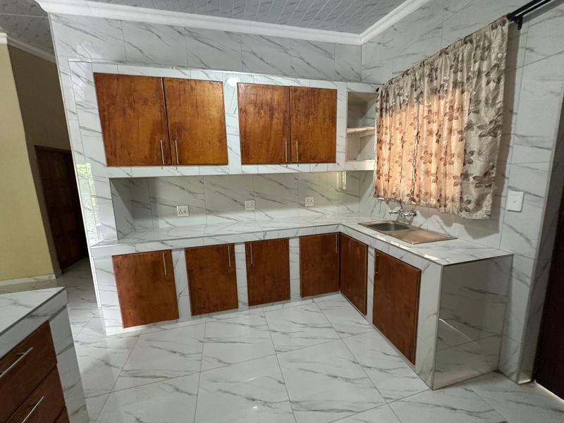 A clean, empty kitchen featuring marble-look tiled walls and countertops, complemented by wooden cabinetry and a stainless steel sink.