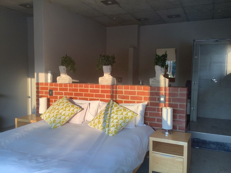 A modern bedroom featuring an exposed brick partition wall, concrete ceiling, and an en-suite shower area with glass enclosure.