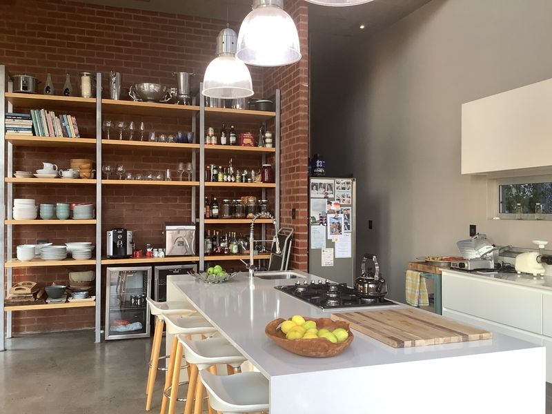 Modern kitchen featuring a large central island with a built-in gas cooktop, stainless steel sink, and open industrial-style shelving against a brick feature wall.
