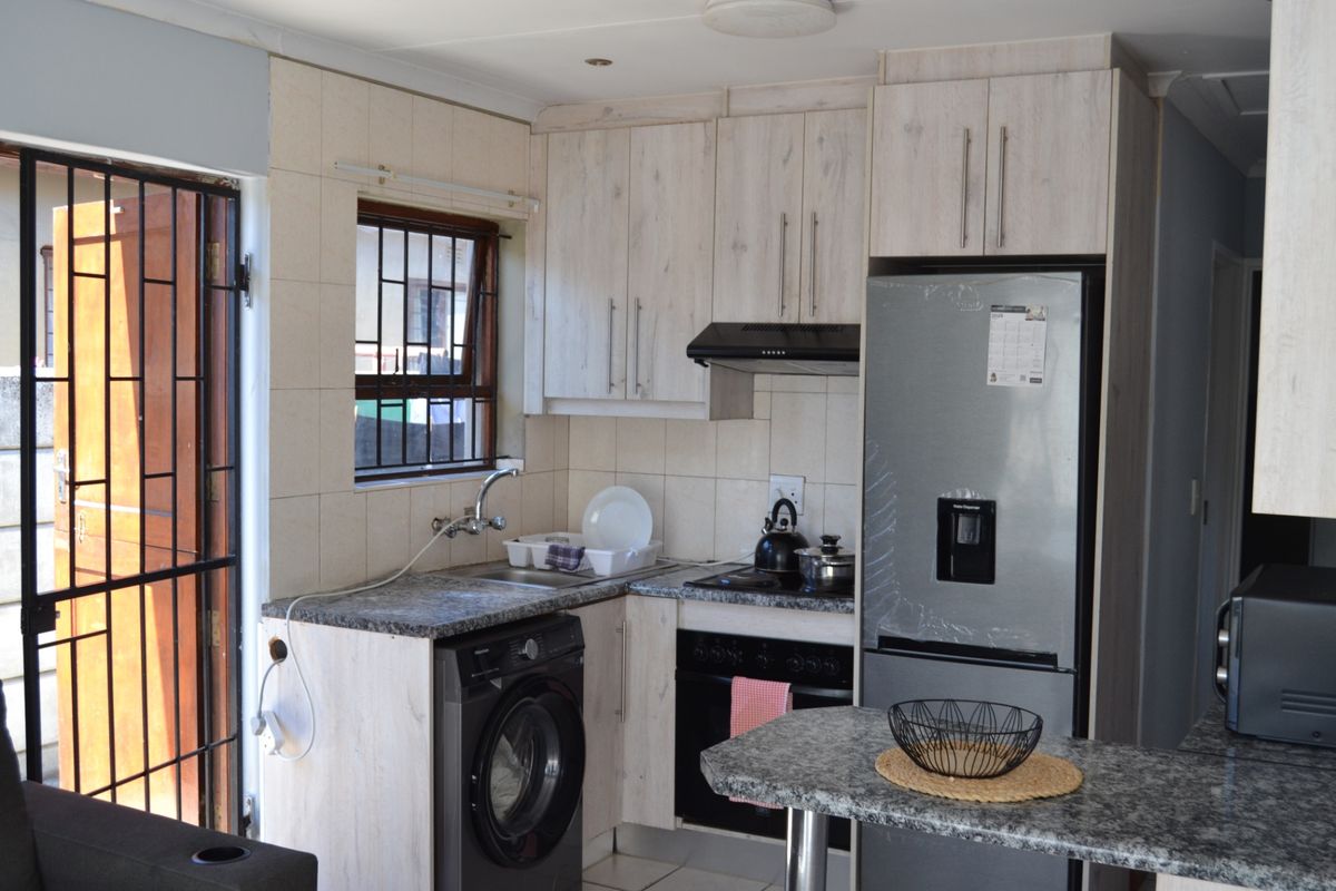 Compact kitchen featuring light wood-look cabinetry, granite countertops, built-in oven and hob, and integrated laundry plumbing, with direct access to an exterior security gate.