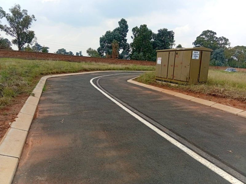 A view of a paved residential street featuring concrete curbing and a prominent electrical transformer box, bordered by a perimeter brick wall.