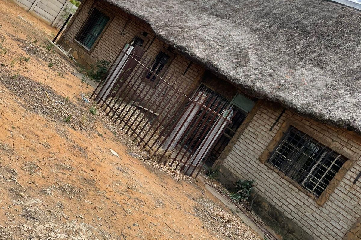 Rear view of a single-story house featuring a traditional thatch roof, exposed brick walls, and metal security bars on windows and doors.