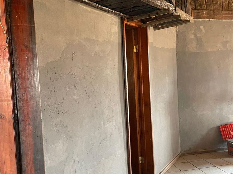 A rustic interior space featuring a thatched ceiling, exposed timber beams, and unfinished plastered walls, leading into an adjacent room through a timber door frame.