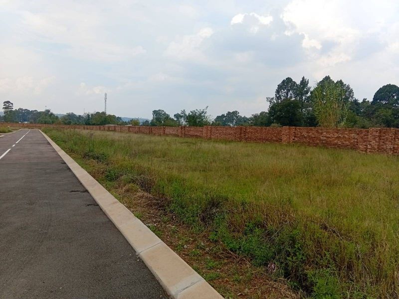 A vacant plot of land featuring a long brick boundary wall, adjacent to a paved road with concrete kerbing.