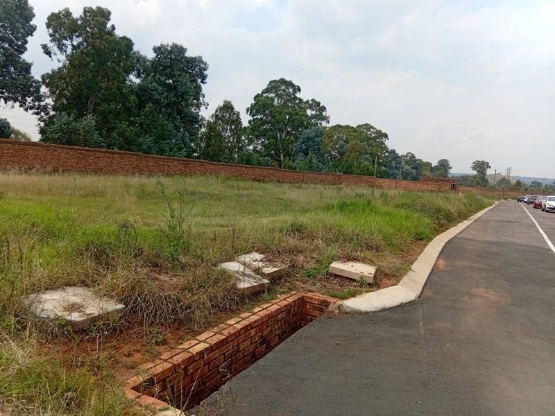 A view of a vacant plot of land featuring a brick boundary wall, asphalt road access, and concrete kerbing.