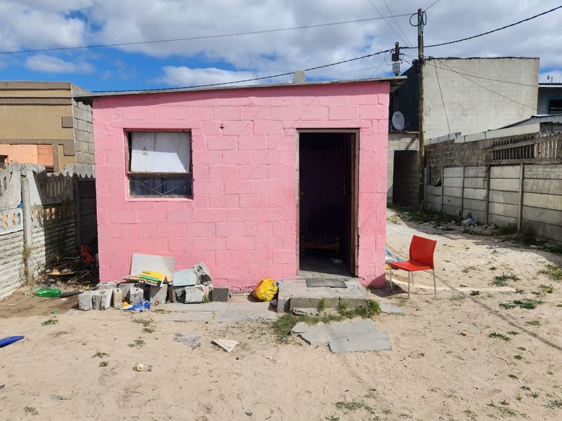 A small, pink-painted concrete block structure featuring a single entrance and window, situated on an unpaved lot with surrounding boundary walls.
