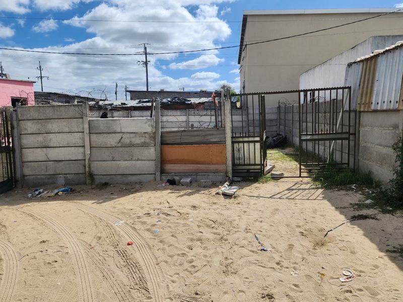 Front exterior view showing a sandy driveway area, metal security gate, and precast concrete boundary wall with visible structural damage and makeshift repairs.