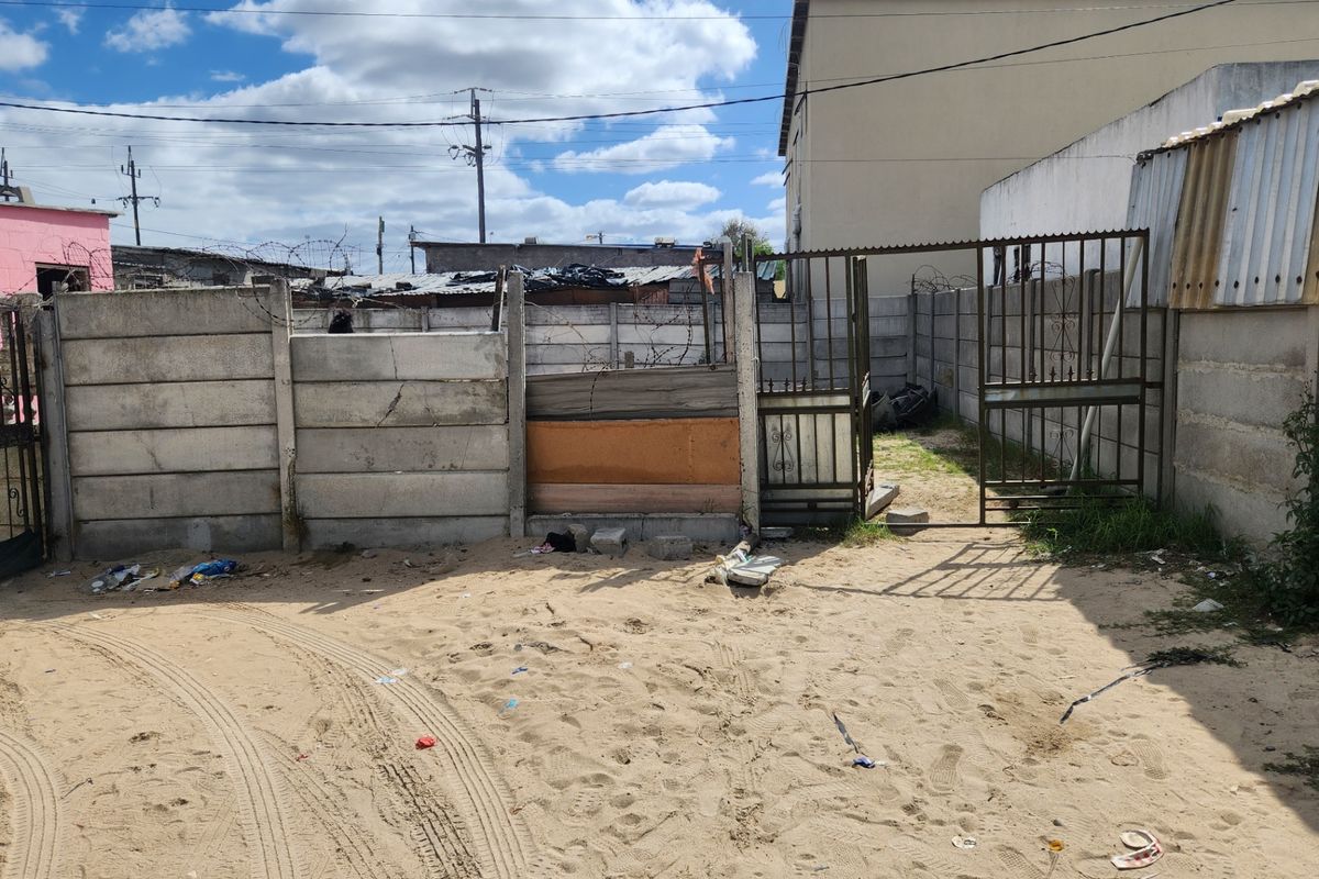 Front exterior view showing a sandy driveway area, metal security gate, and precast concrete boundary wall with visible structural damage and makeshift repairs.