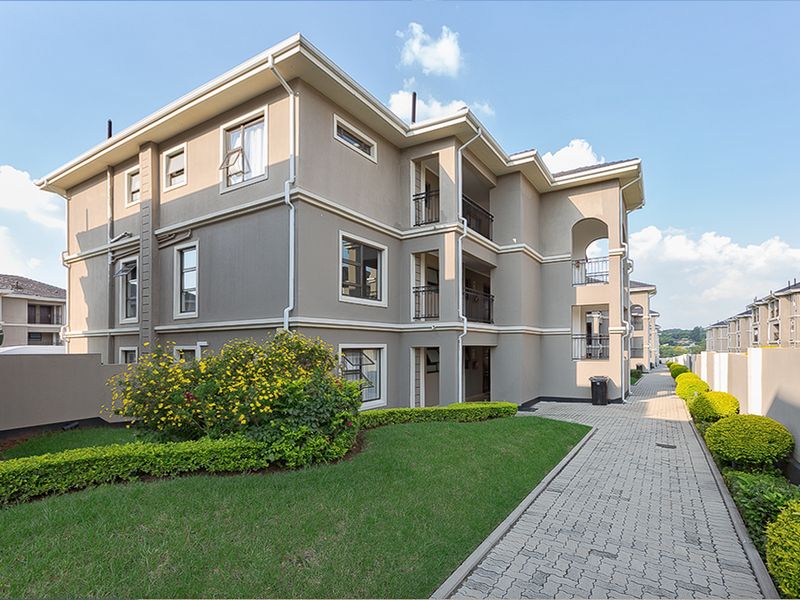 A well-maintained apartment complex exterior featuring a paved walkway, landscaped garden areas, and private balconies with modern architectural finishes.