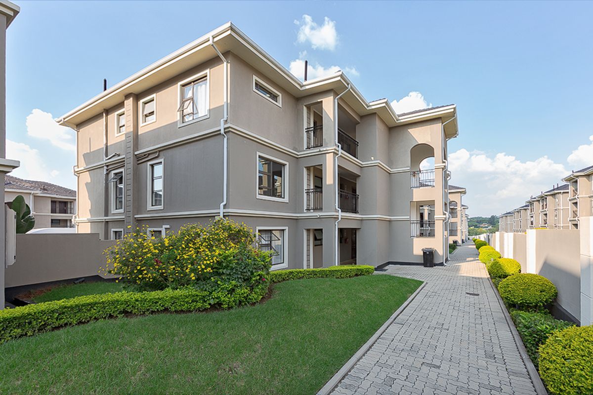 A well-maintained apartment complex exterior featuring a paved walkway, landscaped garden areas, and private balconies with modern architectural finishes.