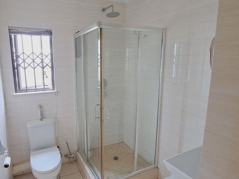 A functional bathroom featuring a glass shower enclosure, toilet, and neutral wall tiling with security bars on the window.