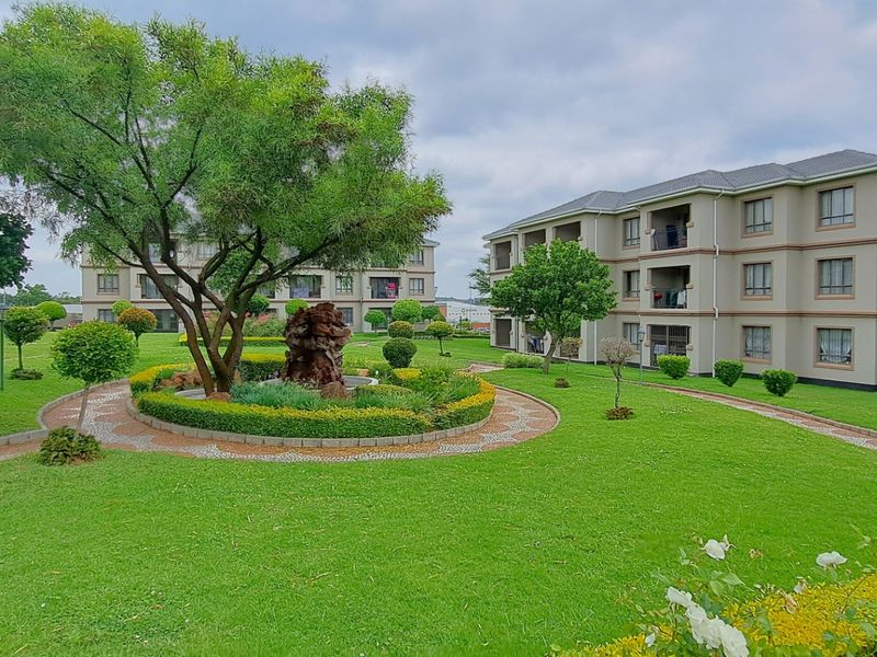 A well-maintained communal garden area featuring manicured lawns, paved walkways, and mature trees situated between multi-story apartment buildings.