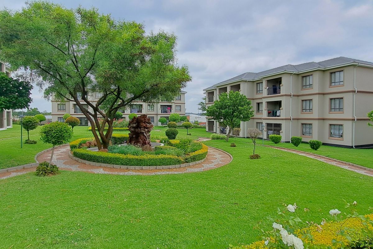 A well-maintained communal garden area featuring manicured lawns, paved walkways, and mature trees situated between multi-story apartment buildings.