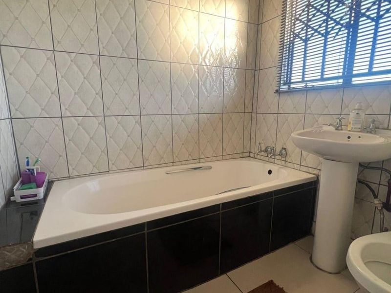 A functional bathroom featuring a bathtub, pedestal basin, toilet, and tiled finishes throughout, complemented by a window with fitted blinds.