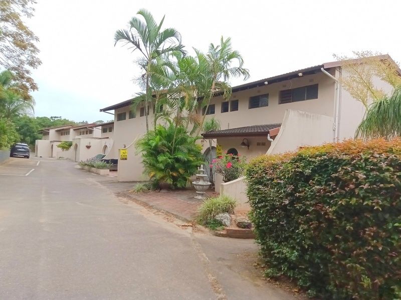 A view of the townhouse complex exterior, featuring a paved driveway, established landscaping, and multi-level residential units with tiled roofing.