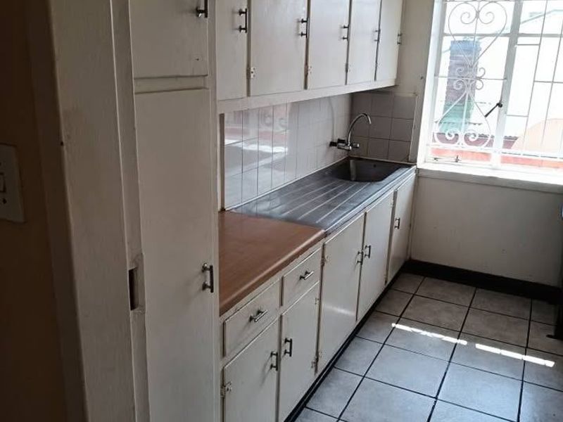 A compact kitchen featuring dated built-in cabinetry, a stainless steel sink, tiled flooring, and a window with a security gate.