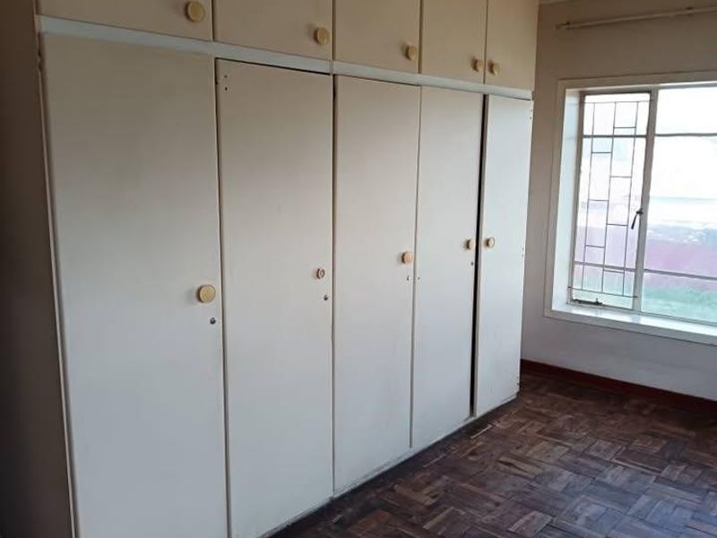 A vacant bedroom featuring extensive built-in cabinetry, classic parquet flooring, and a window fitted with security bars.