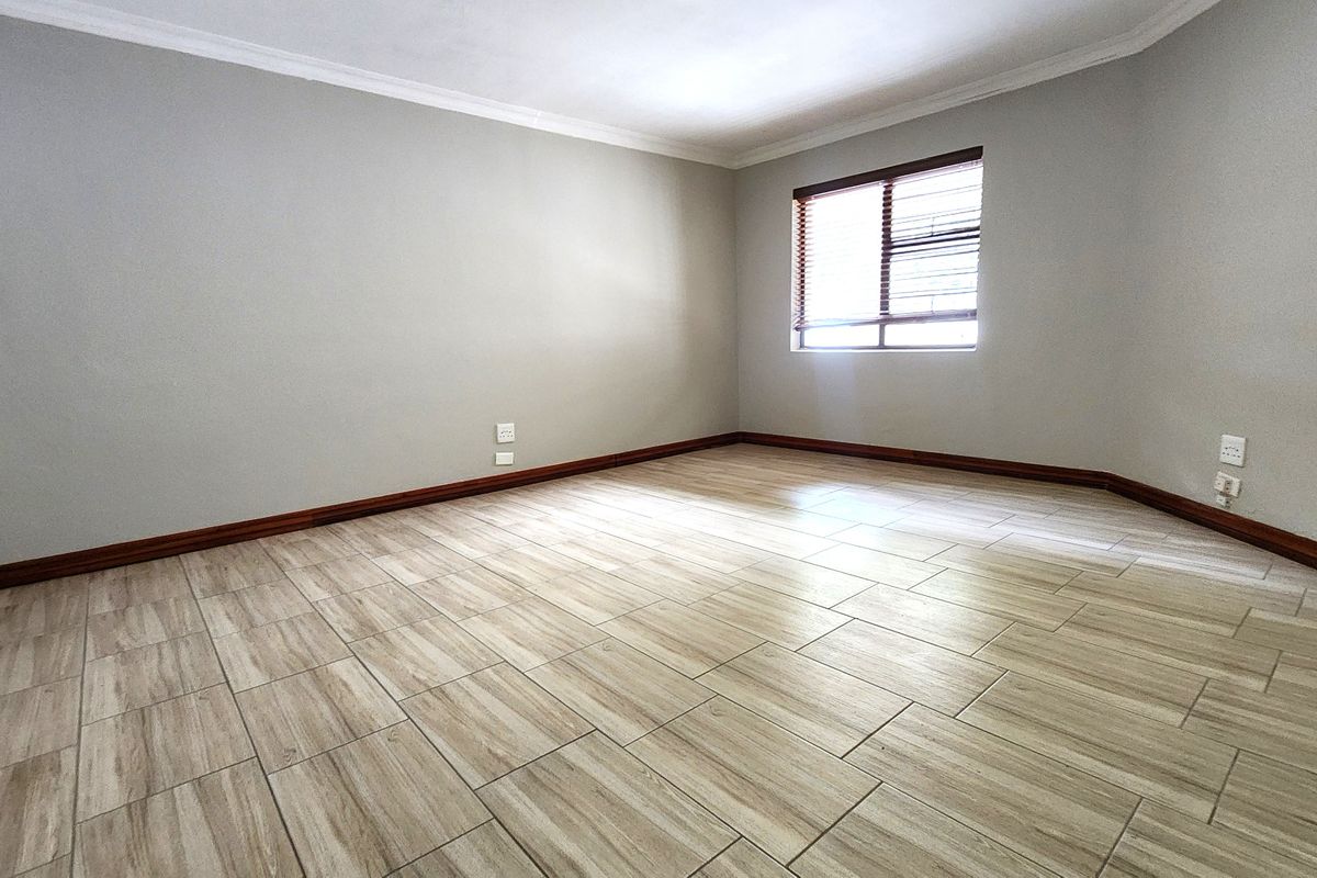 A bright, empty bedroom featuring modern wood-look tiled flooring, neutral grey walls, and fitted venetian blinds.