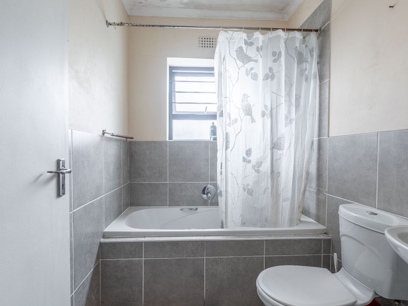 A functional bathroom featuring a bathtub with shower attachment, toilet, and washbasin, finished with grey wall tiles and showing signs of ceiling wear.