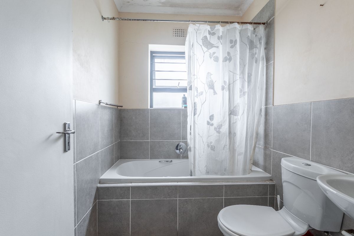 A functional bathroom featuring a bathtub with shower attachment, toilet, and washbasin, finished with grey wall tiles and showing signs of ceiling wear.