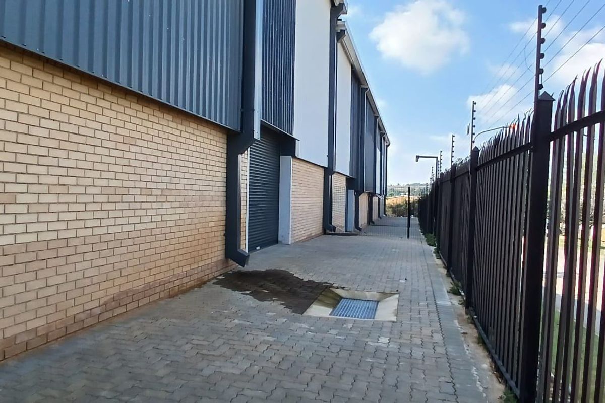 Side exterior view of an industrial property featuring a brick and metal facade, roller shutter door, paved walkway, and secure perimeter fencing.
