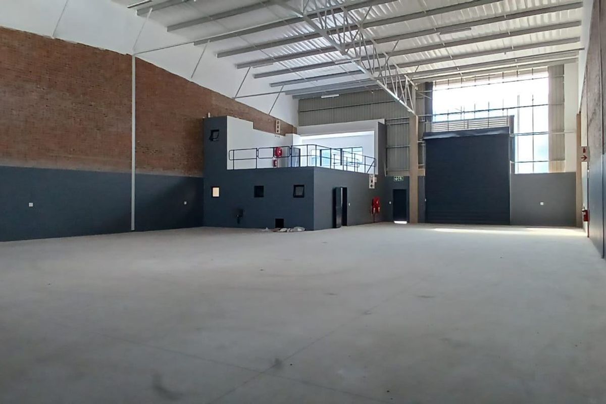 Spacious industrial warehouse unit featuring high ceilings with exposed steel trusses, a mezzanine level, concrete flooring, and a large roller shutter door.