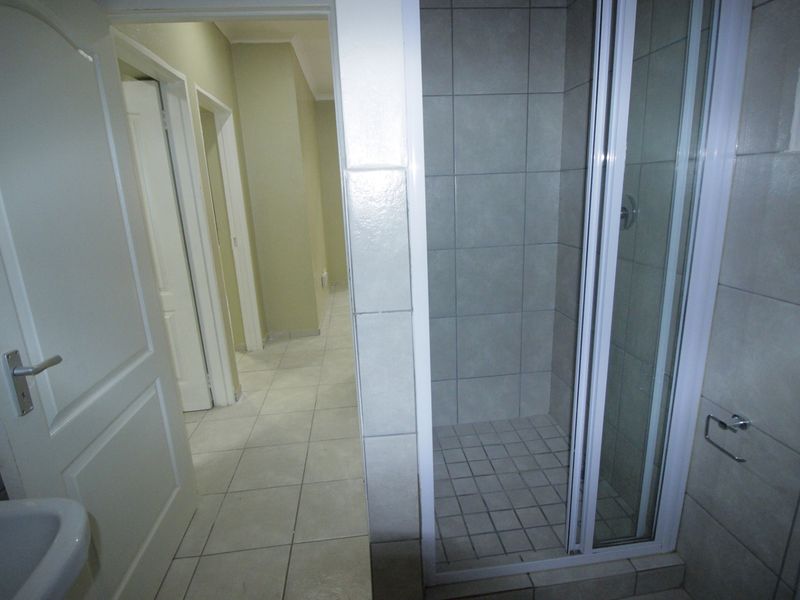 A functional bathroom featuring a tiled shower enclosure, pedestal basin, and neutral floor tiles, with direct access to the hallway.