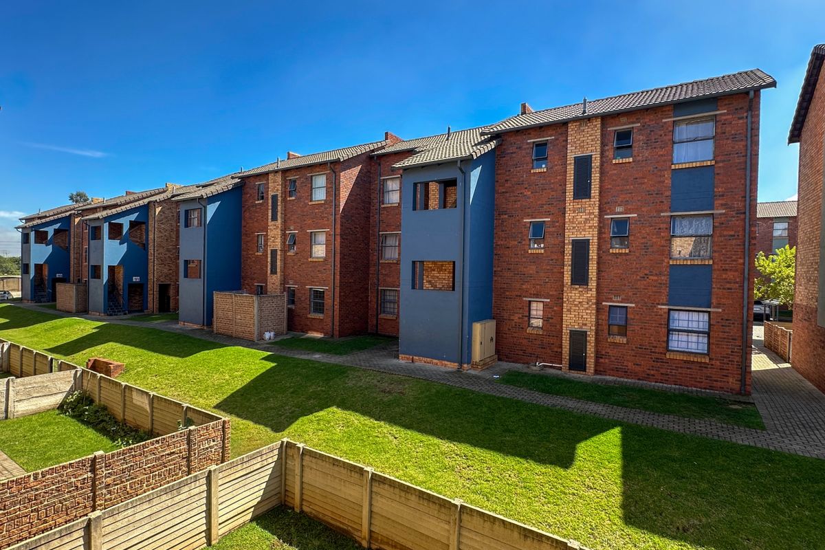 Exterior view of a multi-story brick apartment complex featuring blue accent walls, tiled roofing, and shared green space with paved walkways.