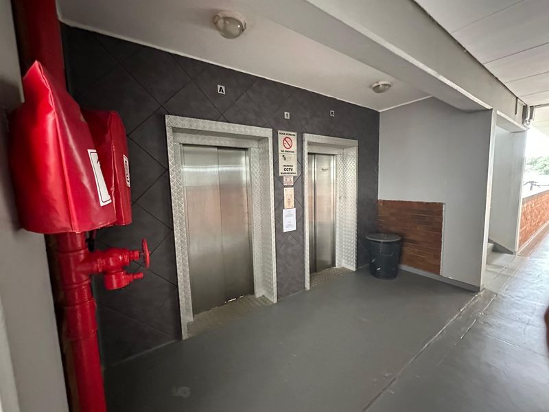 A clean and functional apartment building hallway featuring two stainless steel elevators, a fire hydrant, and modern tiled wall accents.