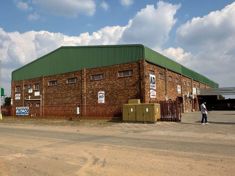A street-facing view of a large industrial warehouse building featuring a brick exterior, green metal roofing, and secure perimeter fencing.