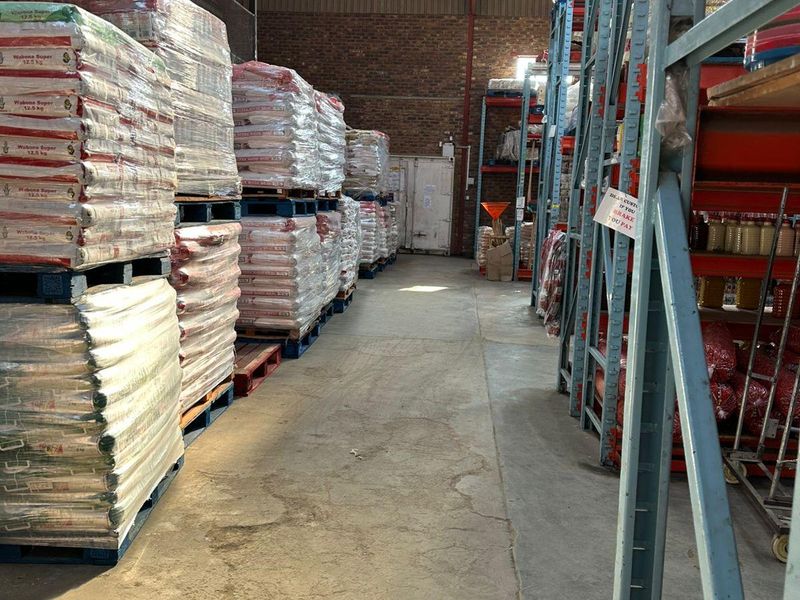 Industrial warehouse space featuring high-ceiling corrugated roofing, skylights, exposed brick walls, and heavy-duty steel racking systems on concrete flooring.