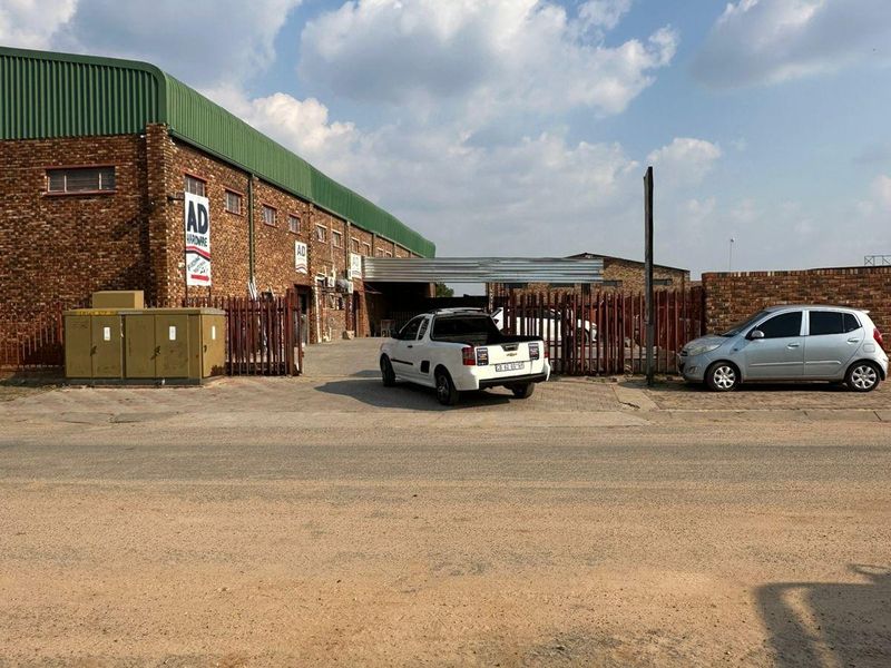 Front exterior view of an industrial warehouse building featuring face brick construction, corrugated metal roofing, and secure perimeter fencing with a paved driveway.