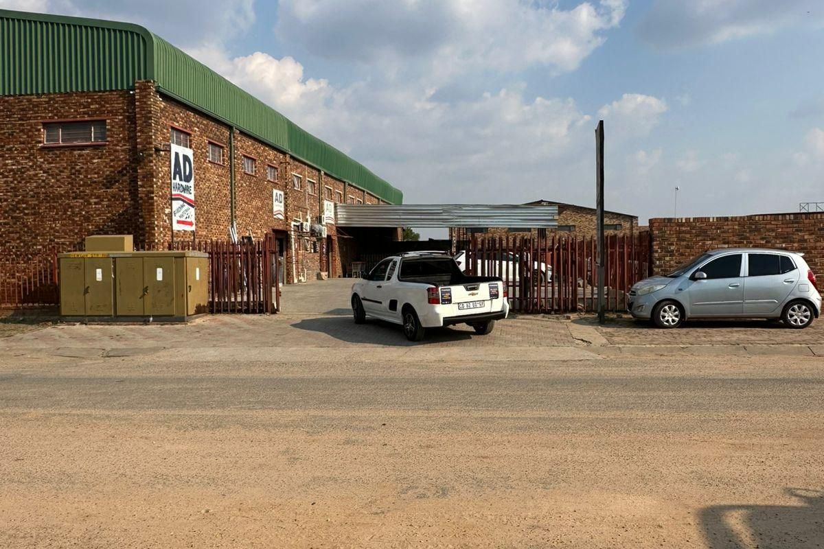Front exterior view of an industrial warehouse building featuring face brick construction, corrugated metal roofing, and secure perimeter fencing with a paved driveway.