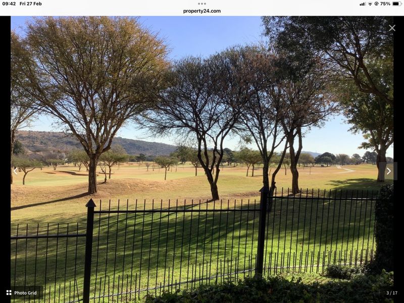 A scenic view overlooking a golf course or open parkland, framed by mature trees and secured by a metal perimeter fence.