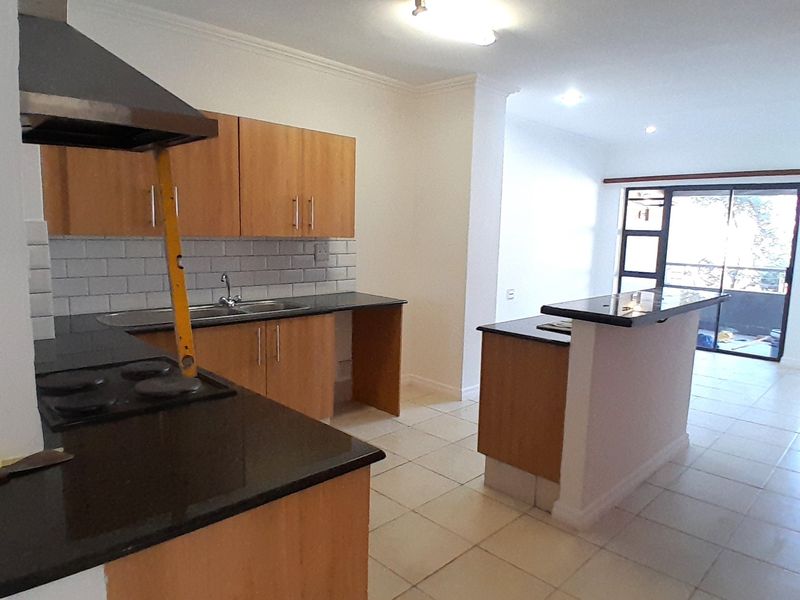 Open-plan kitchen and living area featuring wooden cabinetry, granite countertops, tiled flooring, and a breakfast bar leading to a balcony.