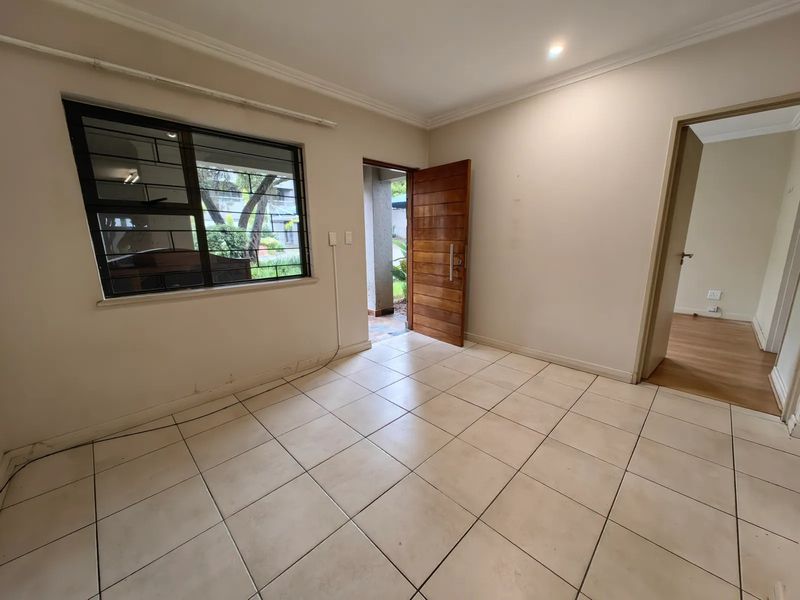 An empty living area featuring tiled flooring, a wooden entrance door, and security bars on the window, offering a blank canvas for personalization.