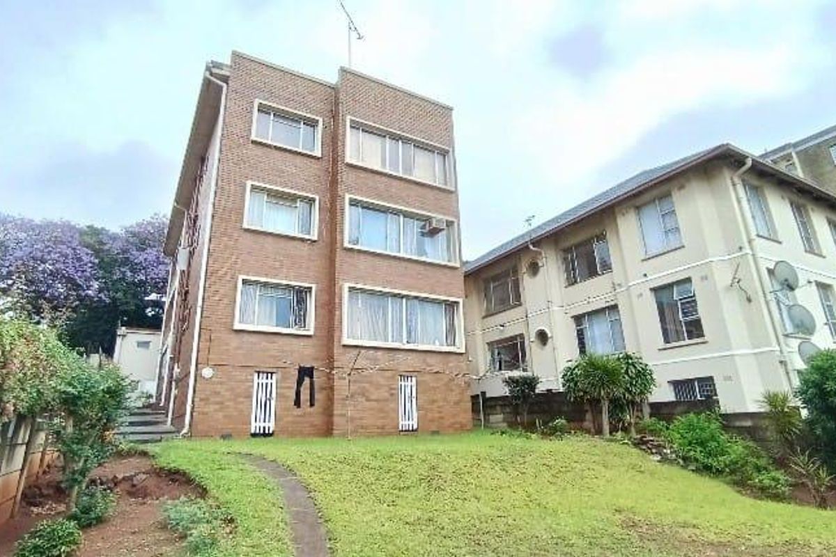 Rear view of a multi-story brick apartment building featuring a shared lawn area and established garden surroundings.