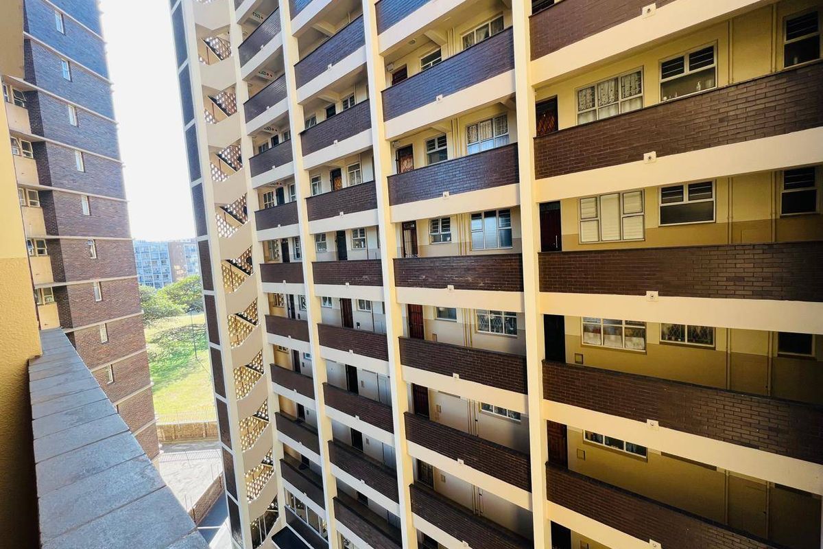 A view of the apartment building exterior featuring brick cladding, multiple levels of balconies with metal railings, and standard window configurations.